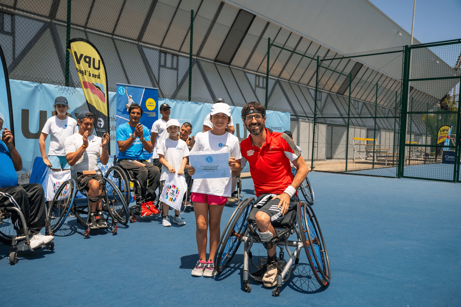 Momento del torneo en pista: participantes, niña con diploma y jugador en silla, UPV al fondo